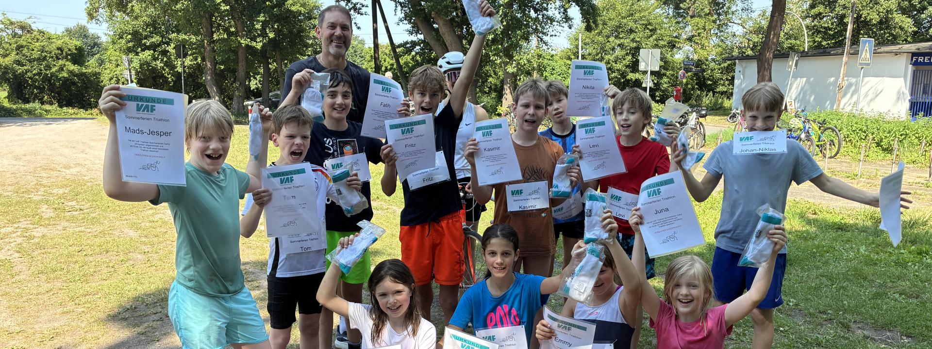 Die 13 Nachwuchsathletinnen und -athleten der Triathlon-Schnupperwoche jubeln mit ihren Urkunden am Freibad, dahinter ihr Betreuer.