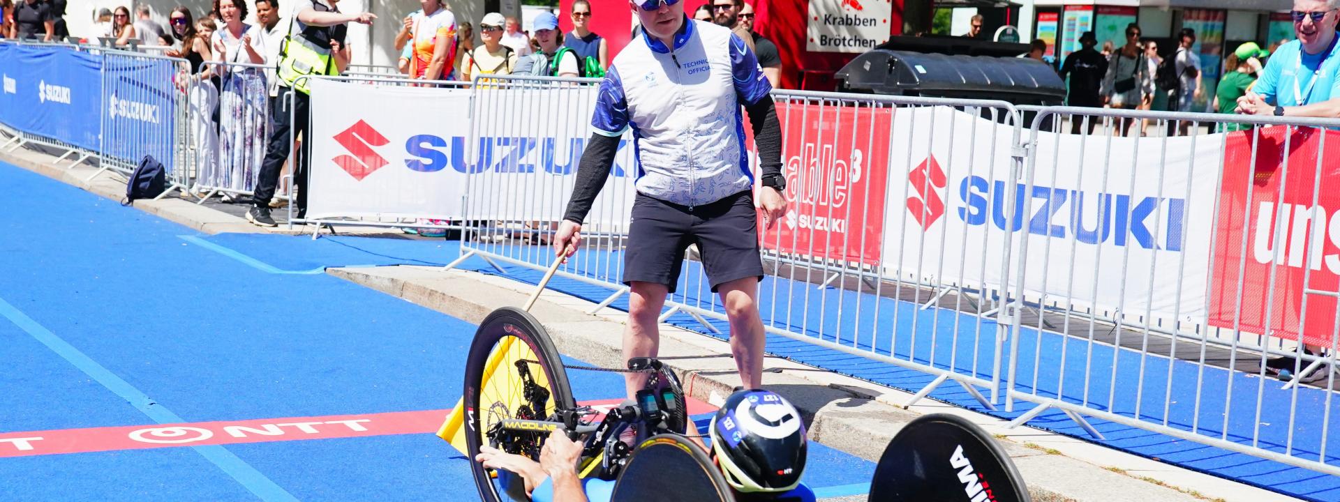 Ein Technical Official (Kampfrichter) dirigiert in der Wechselzone auf dem Jungfernstieg einen Handbiker der PTWC-Klasse.