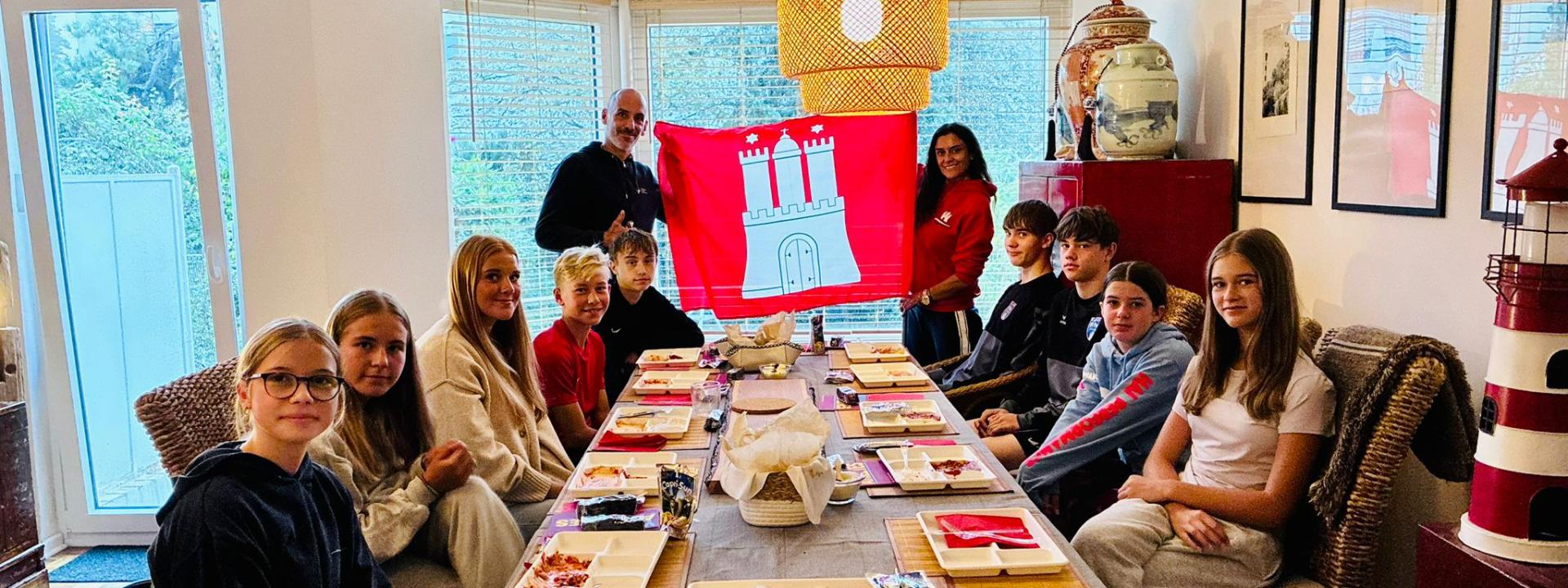 Hamburgs „Jugend trainiert für Olympia"-Team mit Landestrainer und TriAktivistin Loreto Cea beim gemeinsamen Pasta-Essen, im Hintergrund die Hamburg-Flagge.
